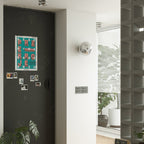 A silver Clérachic wall light on a white wall beside a dark noticeboard with a poster and snapshots, near a glass-block window.