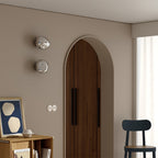 Two silver Clérachic wall lights, stacked on a beige wall beside an arched wooden door and a small black chair.