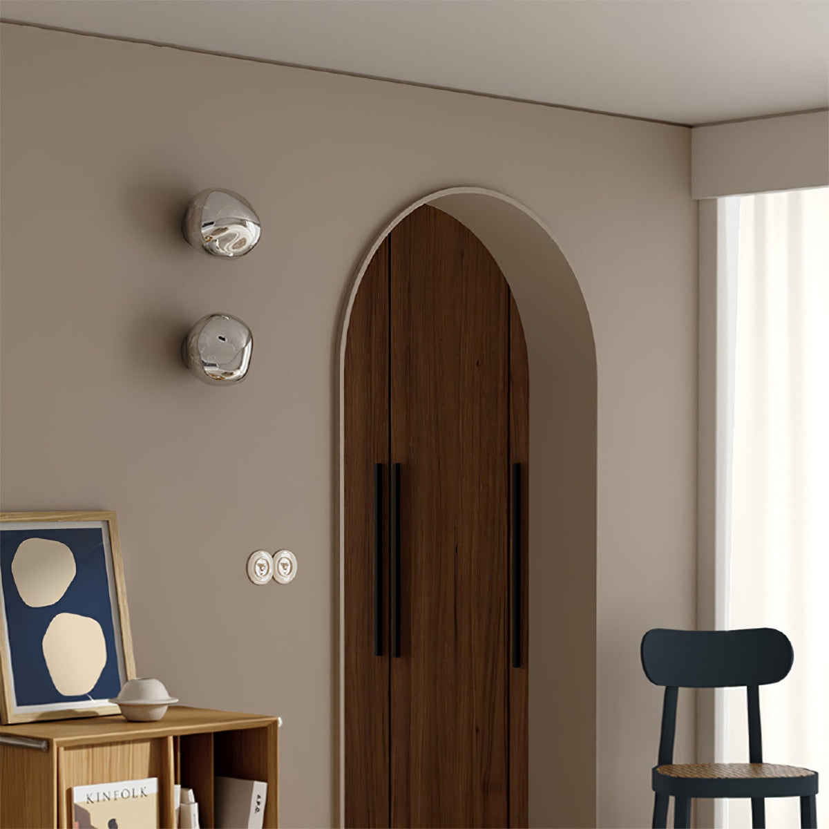 Two silver Clérachic wall lights, stacked on a beige wall beside an arched wooden door and a small black chair.