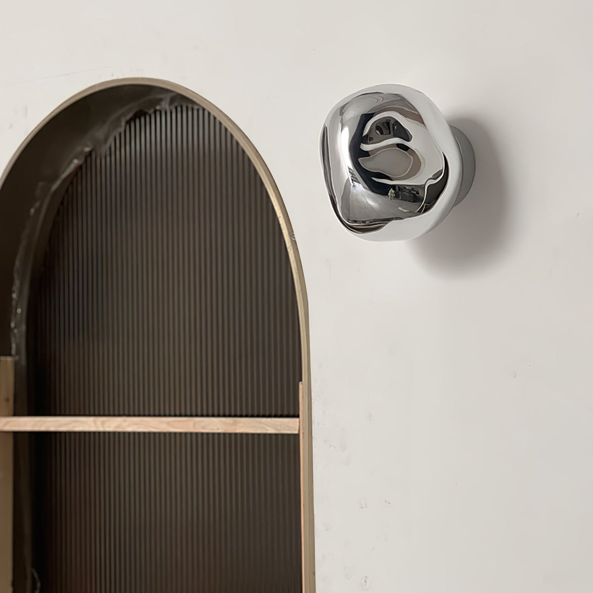A silver, mirror-finish Clérachic wall light on a white wall beside an arched alcove with ribbed panelling.