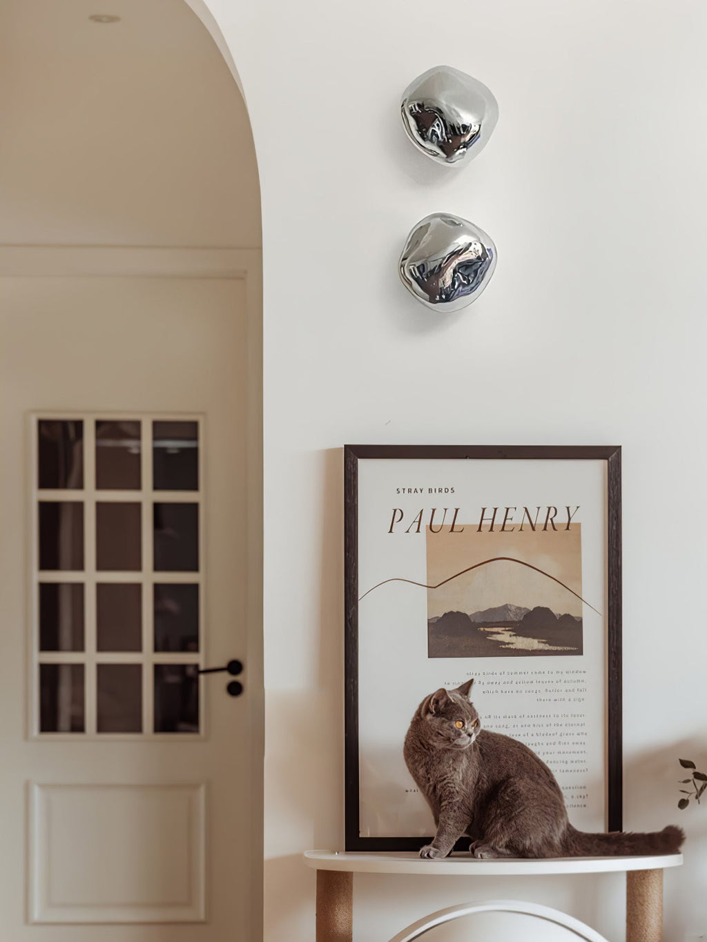 Two silver Clérachic wall lights on a cream wall, with a poster of a cat sitting beneath them in front of a panelled door.