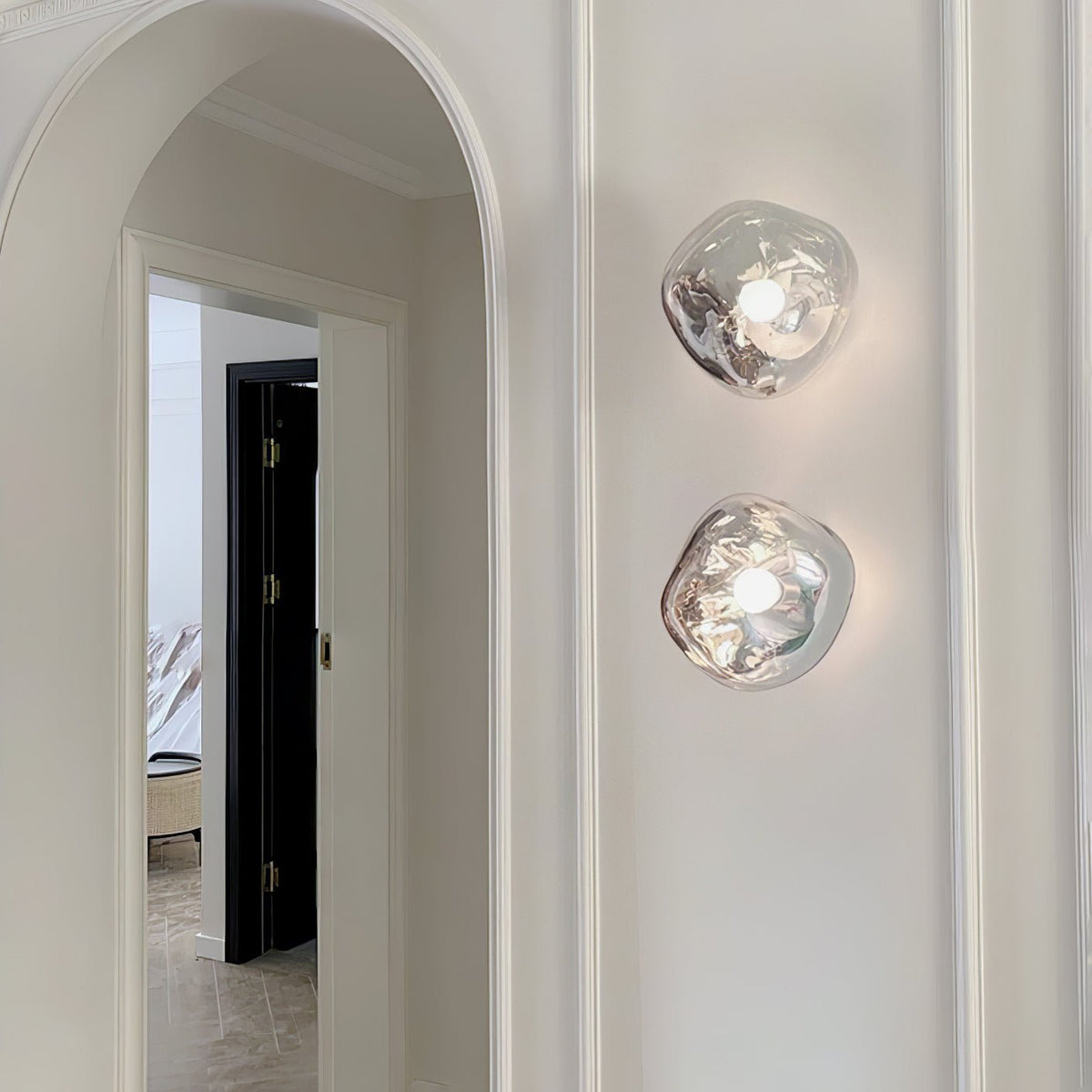 Two Clérachic wall lights with clear glass shades, lit on a white wall beside an arched doorway with a long moulding.