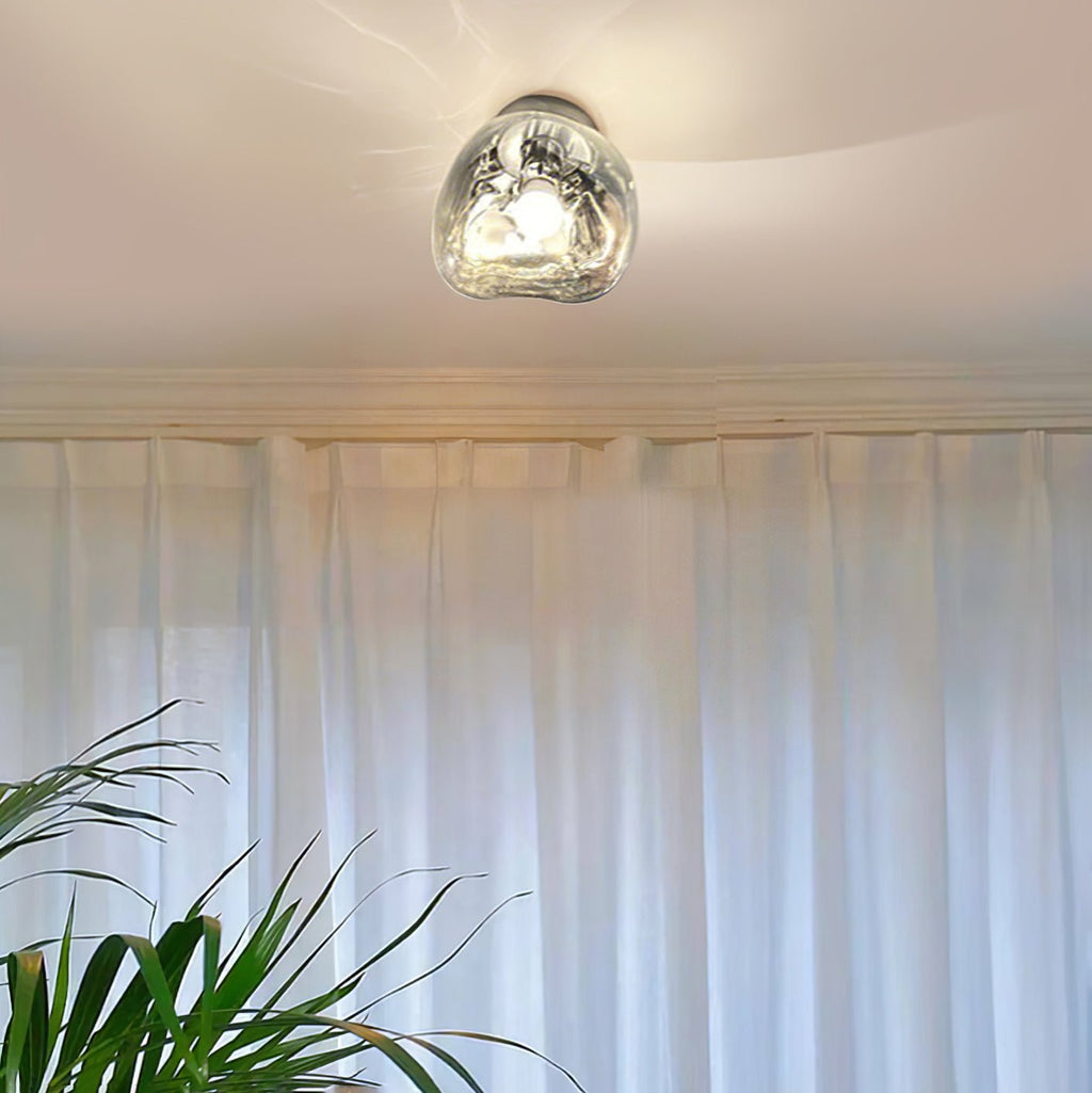 A Clérachic ceiling light with a clear glass shade, lit above sheer white curtains and a leafy plant.