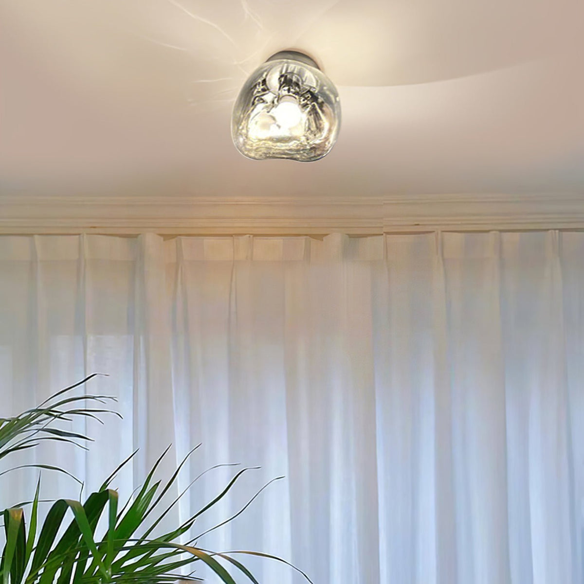 A Clérachic ceiling light with a clear glass shade, lit above sheer white curtains and a leafy plant.