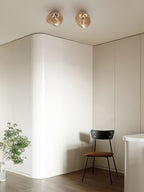 Two Clérachic ceiling lights in a warm champagne tone, above a curved white wall with a chair and a plant below.