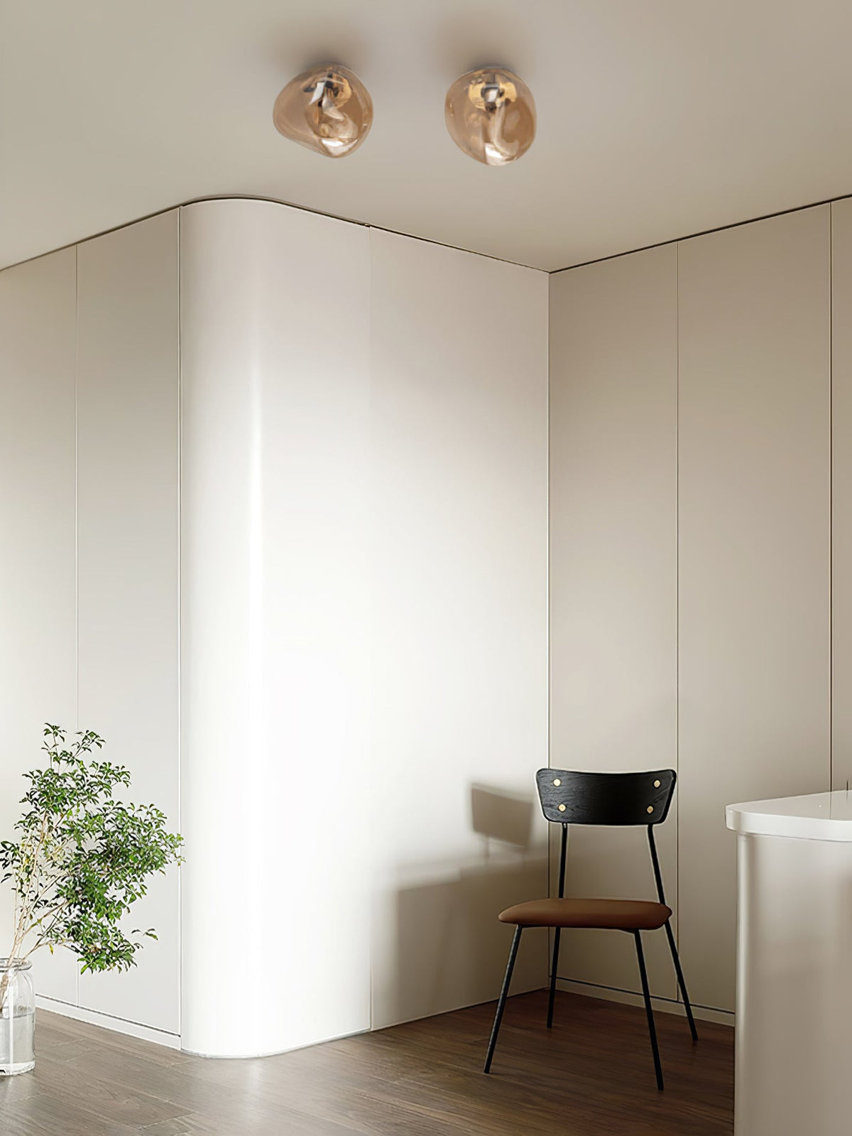 Two Clérachic ceiling lights in a warm champagne tone, above a curved white wall with a chair and a plant below.