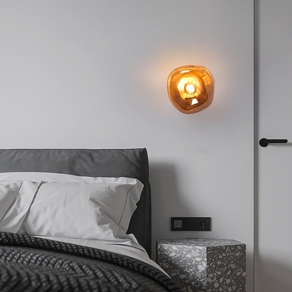 A lit Clérachic wall light with a deep amber glass shade, above a grey bed with dark knit bedding and a terrazzo bedside.