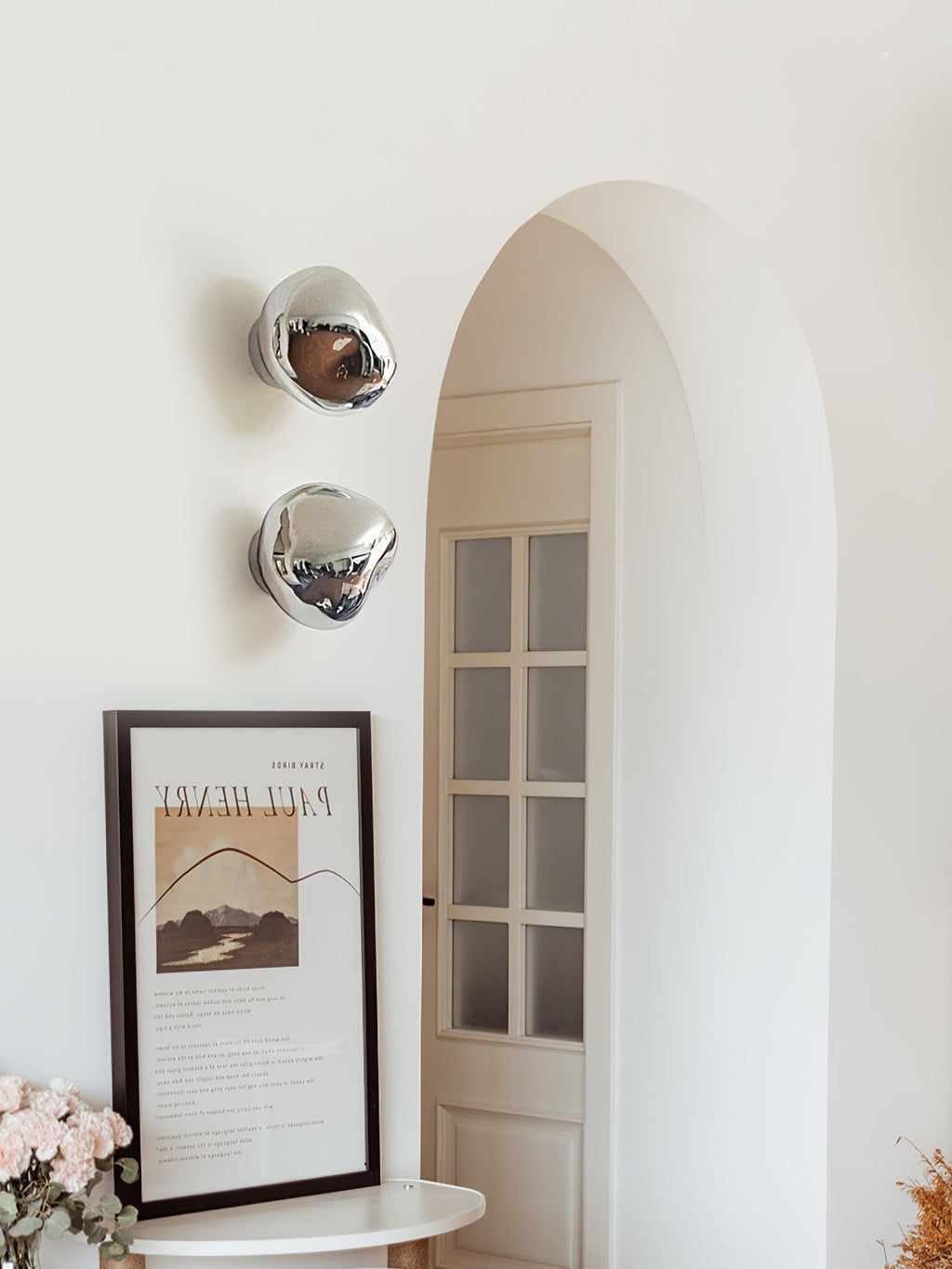 Two silver Clérachic wall lights beside an arched door and a framed print, with fresh flowers at the lower left.