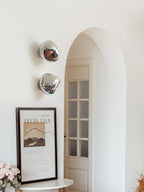 Two silver Clérachic wall lights beside an arched door and a framed print, with fresh flowers at the lower left.