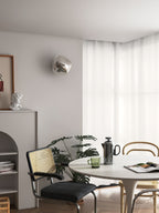 A silver Clérachic wall light on a grey wall above a dining corner with a round table, black chair and sheer curtains.