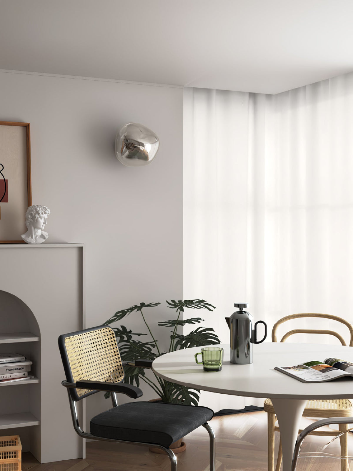 A silver Clérachic wall light on a grey wall above a dining corner with a round table, black chair and sheer curtains.