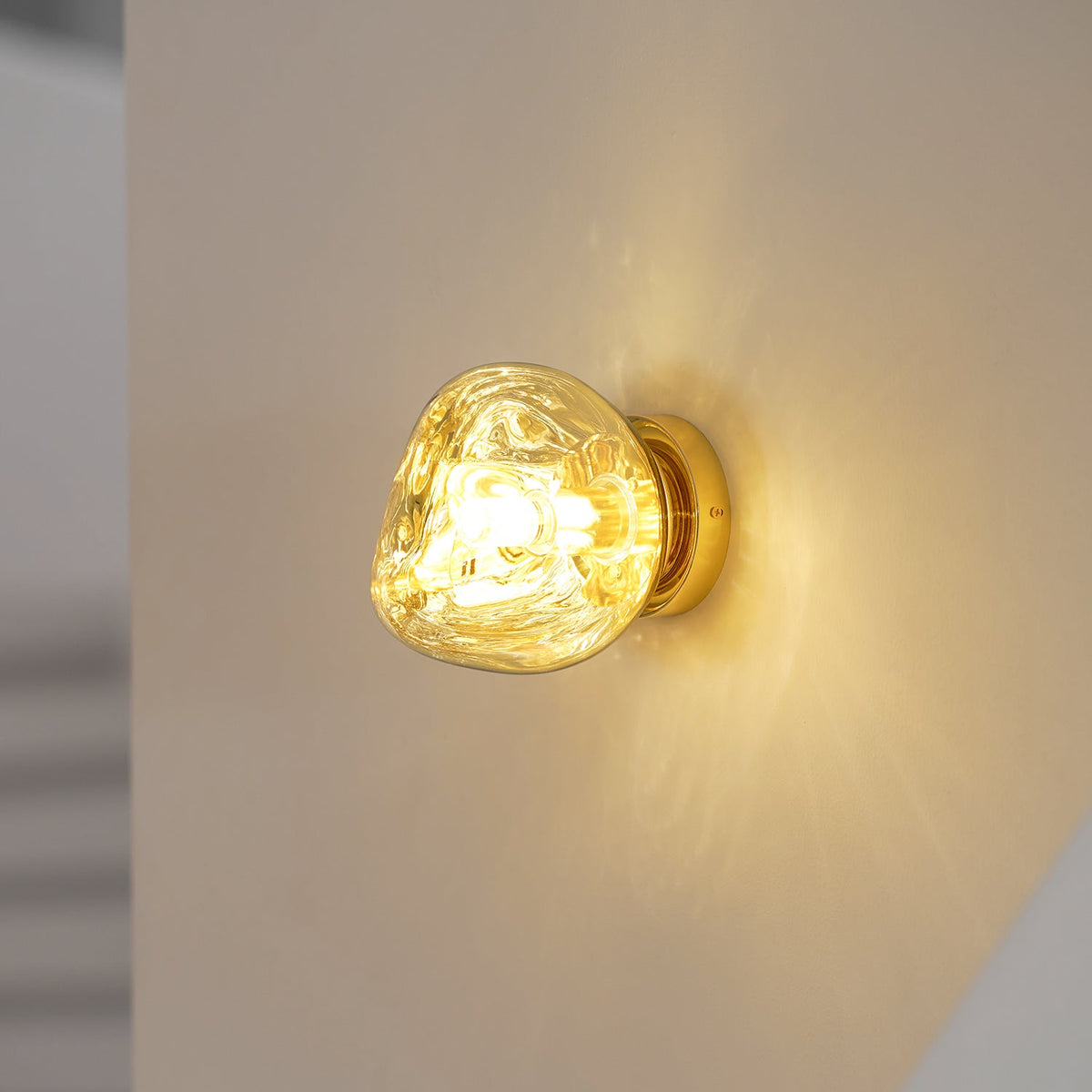Close view of a gold Clérachic wall light glowing warm on a cream wall, with light patterns spreading from the shade.