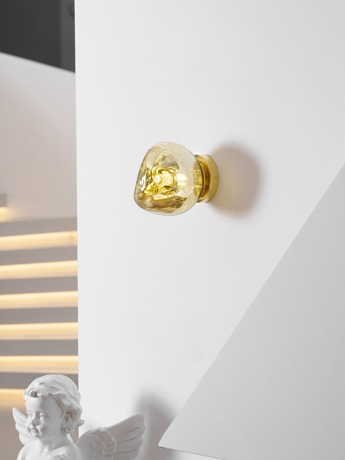 A small gold Clérachic wall light on a white wall, with a cherub figurine and a stepped light strip beyond.