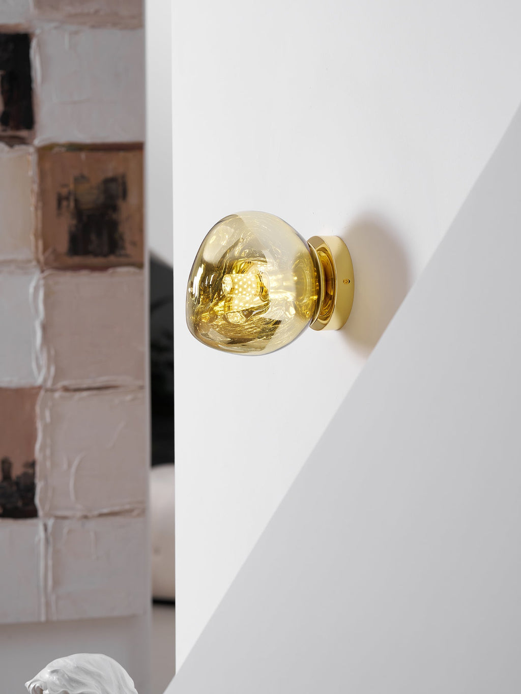 A lit gold Clérachic wall light on a white wall corner, next to a textured painted-brick panel.