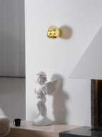 A gold Clérachic wall light on a tall white wall, above a cherub statue on a low plinth and a black mug.
