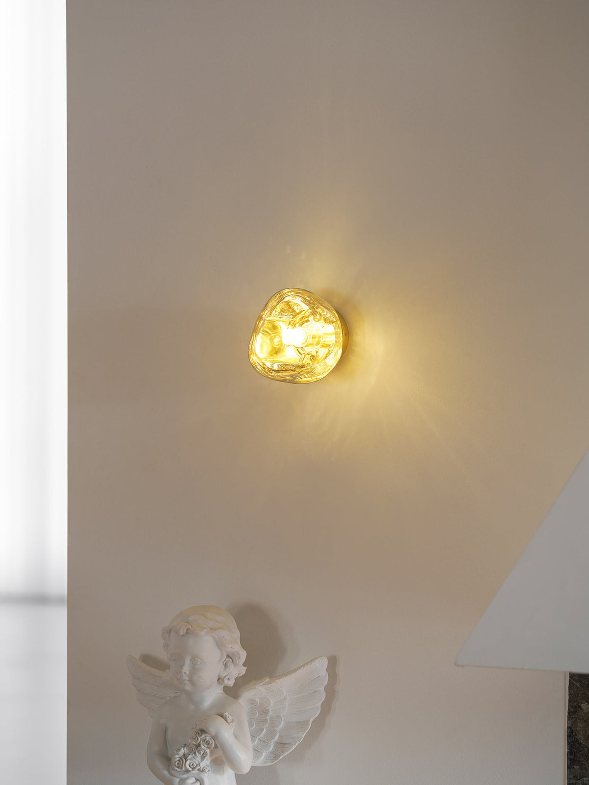 A lit gold Clérachic wall light on a white wall with a cherub figure standing below and a stone surround to the right.