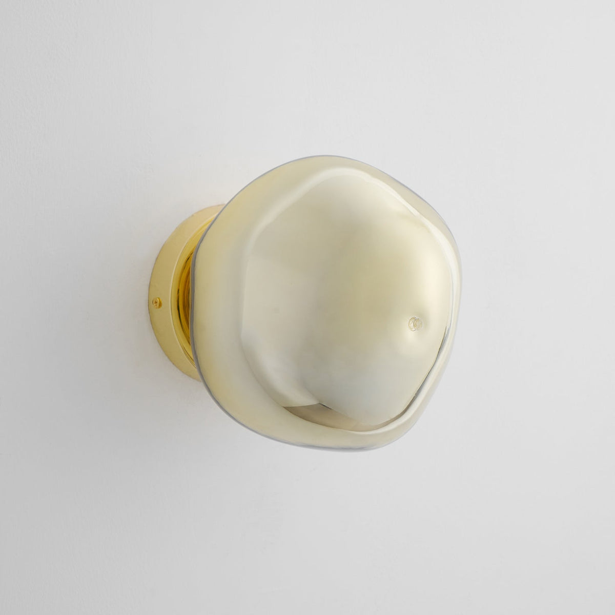A gold Clérachic wall light with a smooth, pale glass shade, unlit, on a pale grey wall with a gold backplate.