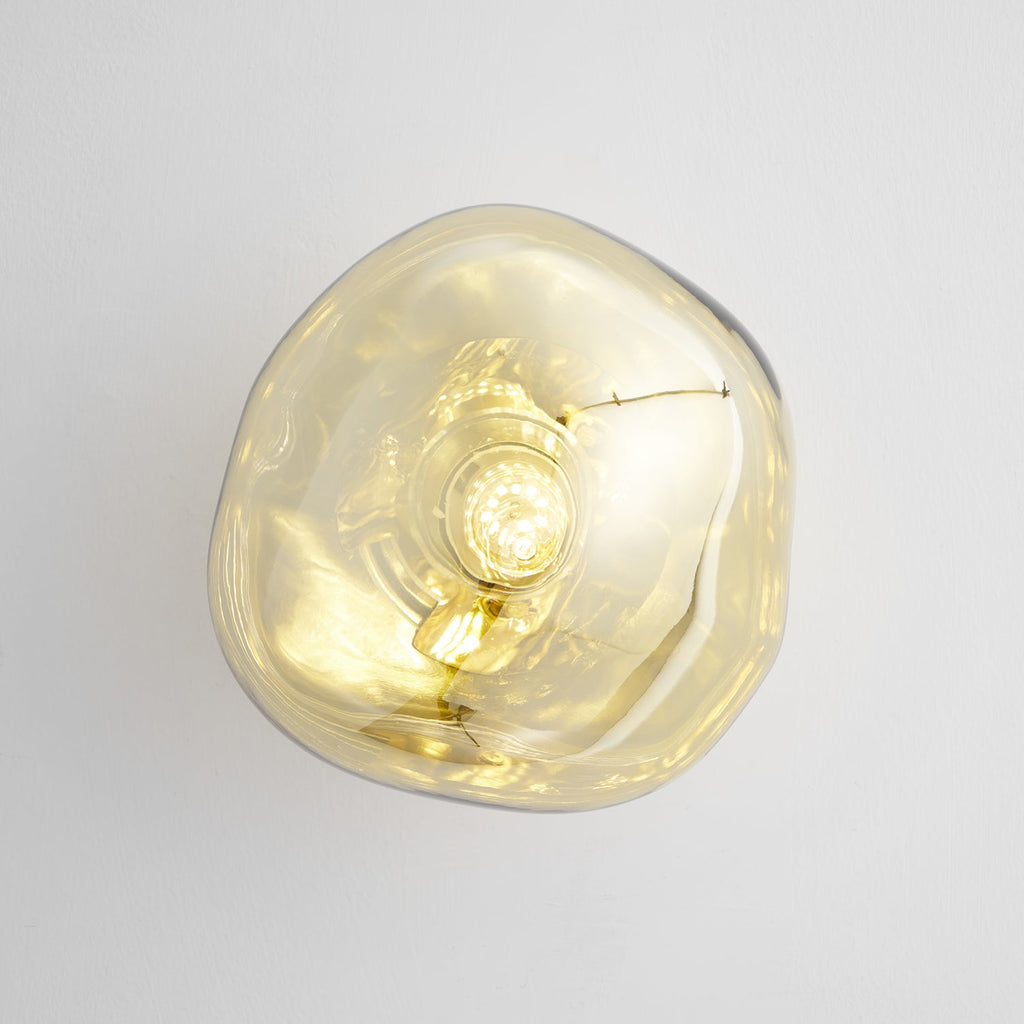 Close view of a gold Clérachic wall light with a clear melted-glass shade, with the bulb showing inside, on a pale wall.