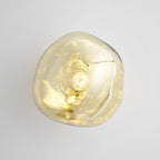 Close view of a gold Clérachic wall light with a clear melted-glass shade, with the bulb showing inside, on a pale wall.