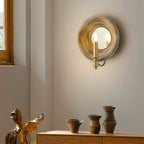 A Clérachic round wood wall light with a candle bulb, on a pale wall beside a window, above a low cabinet with stoneware pots.