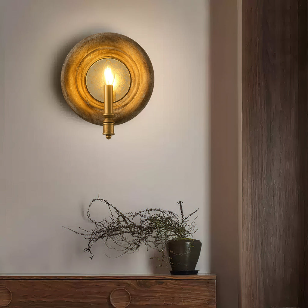 Lit Clérachic wall light with a round wood surround and a gold candle fitting, above a wooden sideboard and a potted plant.