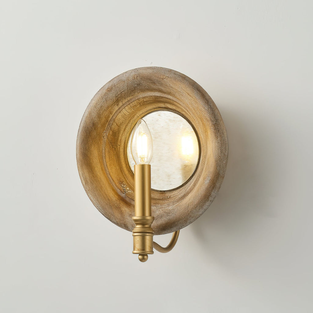 Angled view of a Clérachic wall light with a round wood surround, a mirrored disc and a gold candle-style arm, on a pale wall.