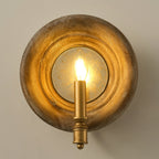 A Clérachic round wood wall light, lit, with a warm glow filling its mirrored centre, on a taupe wall.