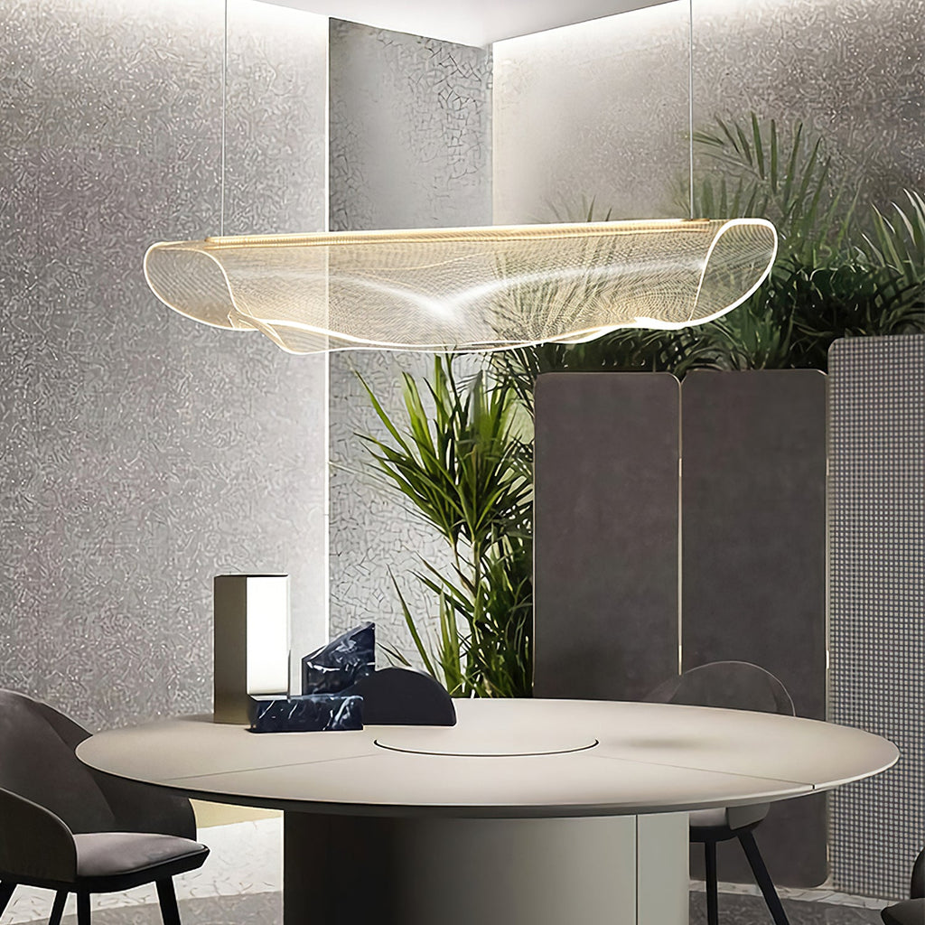 A Clérachic pendant light with its shade curled up at the ends hangs over a round table in a room with tall green plants.