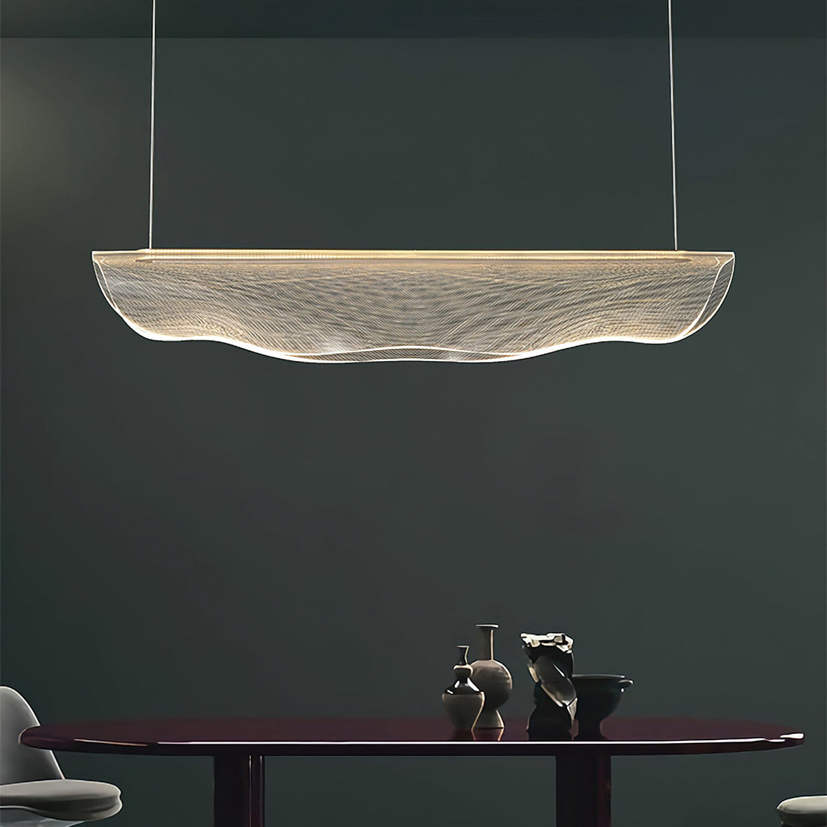 In a dark green-grey room, this Clérachic pendant light hangs above a dark oval table with small ceramic pieces.