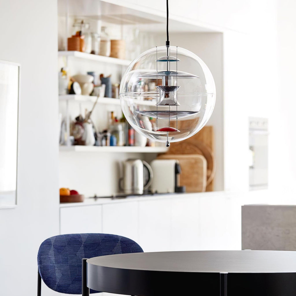 A Clérachic acrylic sphere pendant hangs in a bright kitchen-dining area, with open shelves behind it and a navy chair at a dark round table.