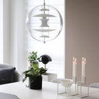 Against a window with blinds, this Clérachic sphere pendant shows white metal discs inside clear acrylic, above a table with candles and a plant.