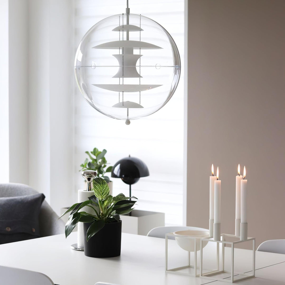 Against a window with blinds, this Clérachic sphere pendant shows white metal discs inside clear acrylic, above a table with candles and a plant.