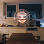 Lit in the evening, this Clérachic sphere pendant casts a soft glow over a dark wood dining table with a kitchen behind it.