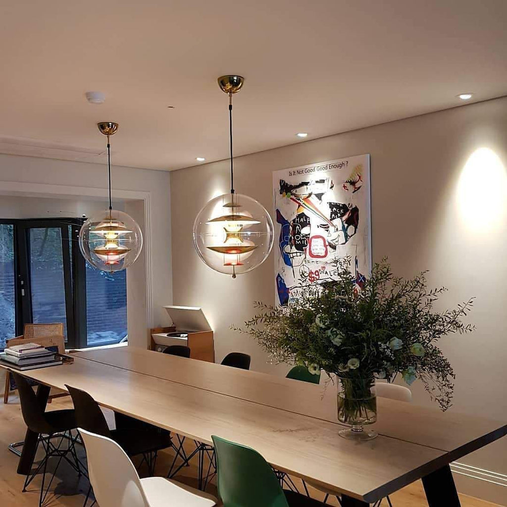 Two Clérachic sphere pendants of different heights hang over a long timber dining table in a room with downlights and a large flower arrangement.