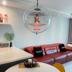 A Clérachic sphere pendant hangs by a pale wall above a round table, with a red leather sofa and a potted palm beyond.