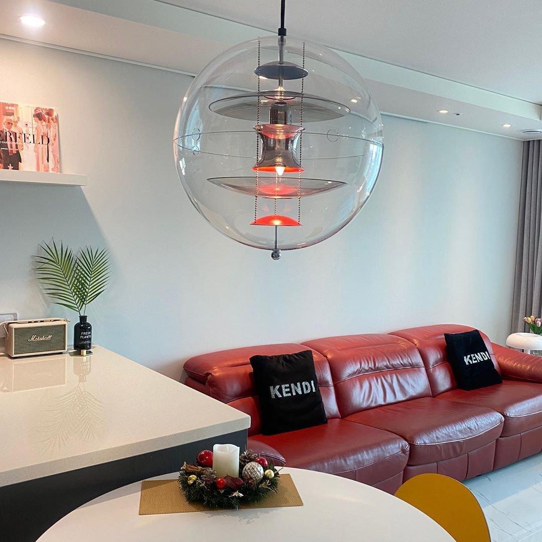 A Clérachic sphere pendant hangs by a pale wall above a round table, with a red leather sofa and a potted palm beyond.