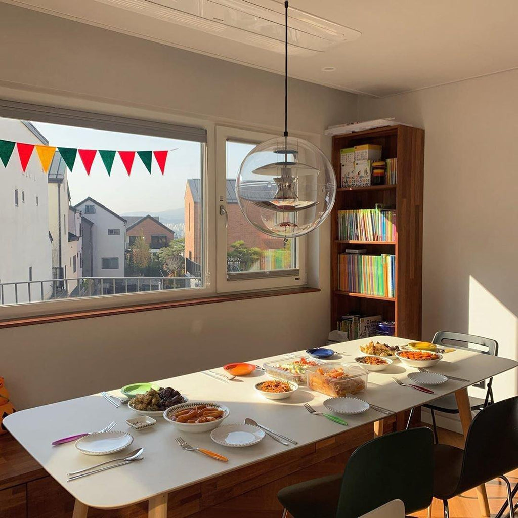A Clérachic sphere pendant hangs by a sunny window over a white table laid with bowls and plates, with a bookcase to one side.