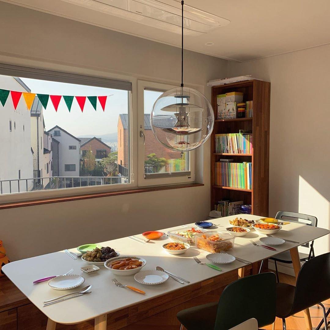 A Clérachic sphere pendant hangs by a sunny window over a white table laid with bowls and plates, with a bookcase to one side.