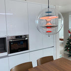 Glowing with a soft orange light, this Clérachic sphere pendant hangs in a white fitted kitchen above a wooden dining table.