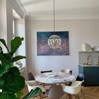 Hung low above a round patterned table, this Clérachic sphere pendant sits in a calm room with a large plant and a painting behind it.
