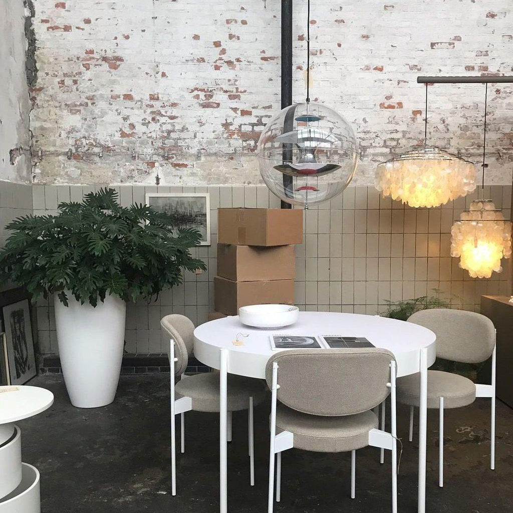A Clérachic sphere pendant hangs beside two shell-style pendants over a white round table in a room with a whitewashed brick wall.