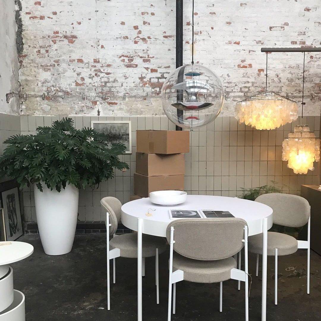 A Clérachic sphere pendant hangs beside two shell-style pendants over a white round table in a room with a whitewashed brick wall.