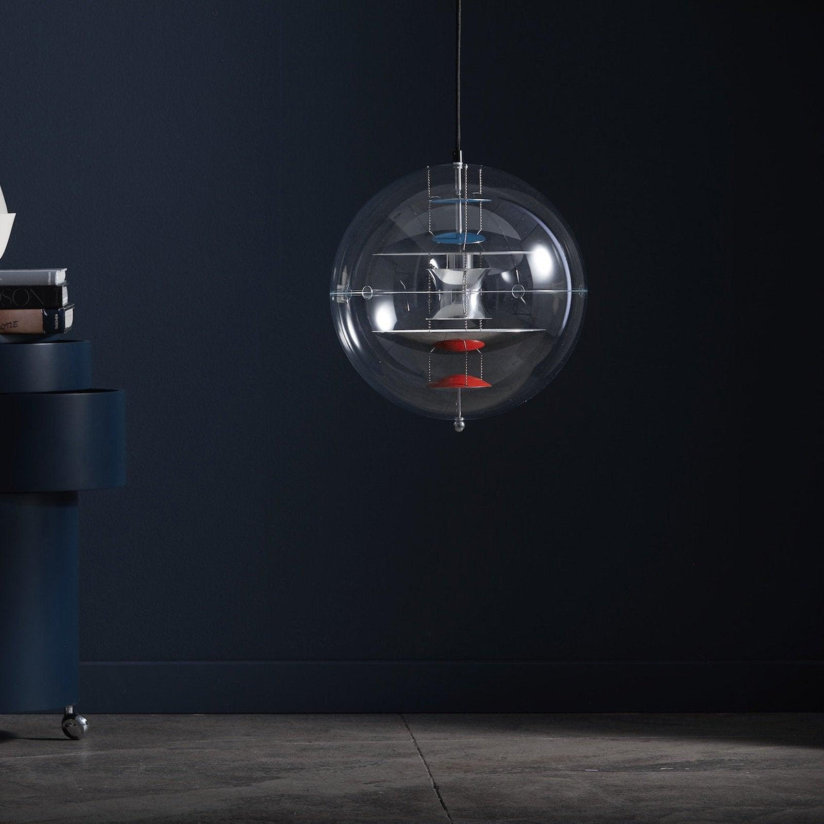 Against a deep navy wall, this Clérachic sphere pendant hangs on a thin cord, its white discs and red dish visible through the clear acrylic.
