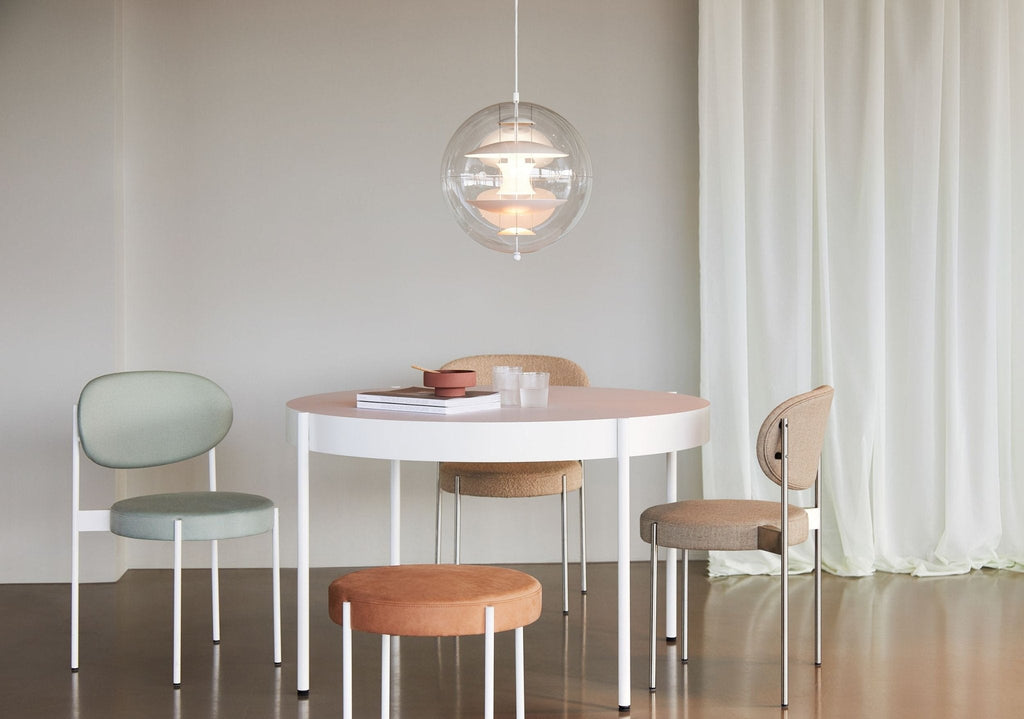 A Clérachic sphere pendant glows warmly above a round pink-topped table with pastel chairs and stools, beside sheer white curtains.