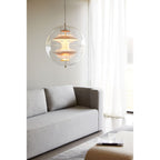A lit Clérachic sphere pendant hangs by white curtains, with peach-toned discs inside, above a grey sofa and a cream ottoman.