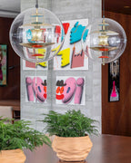 Two Clérachic sphere pendants with gold-toned discs hang in front of a concrete wall hung with bright pop-art prints, above potted ferns.