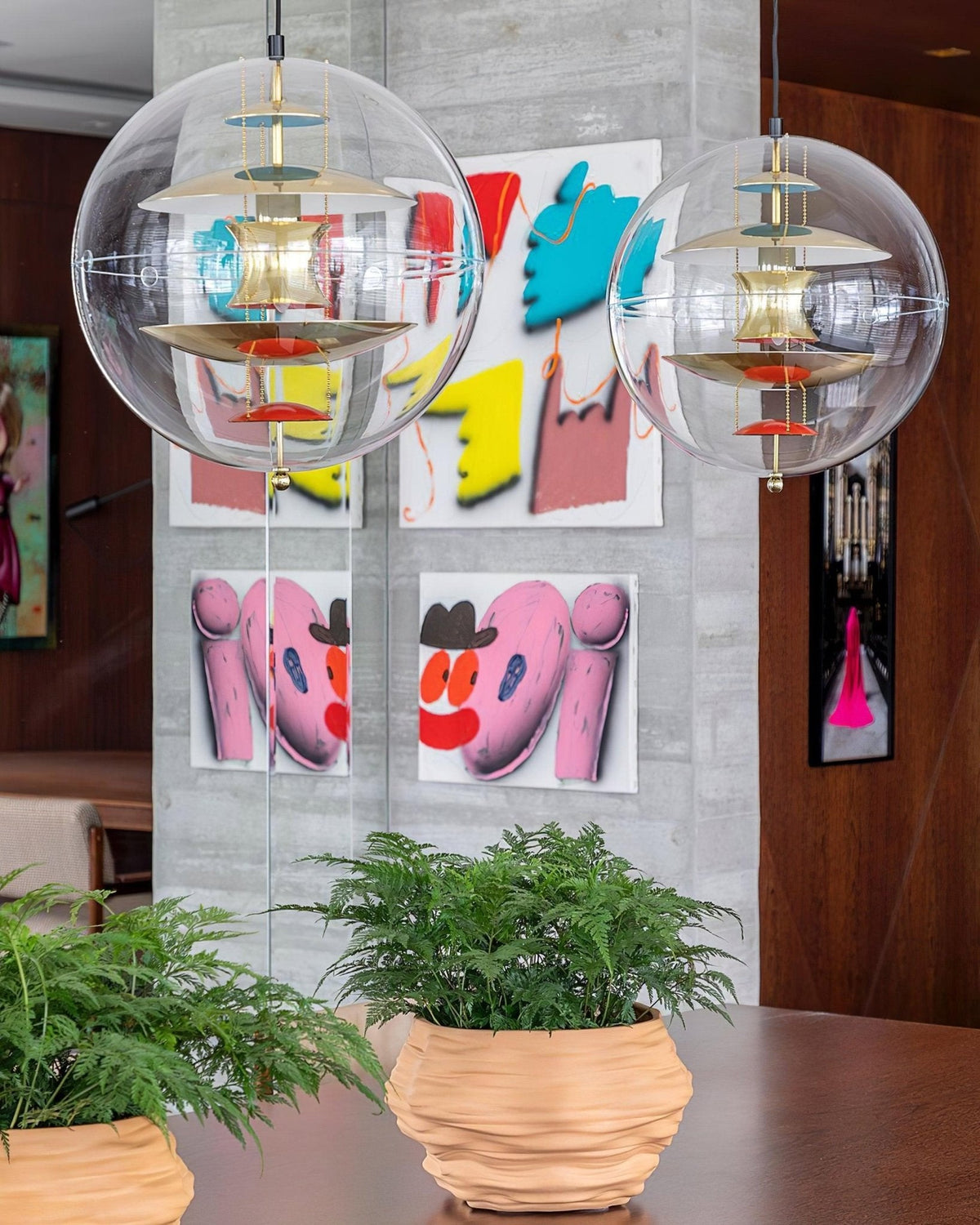 Two Clérachic sphere pendants with gold-toned discs hang in front of a concrete wall hung with bright pop-art prints, above potted ferns.