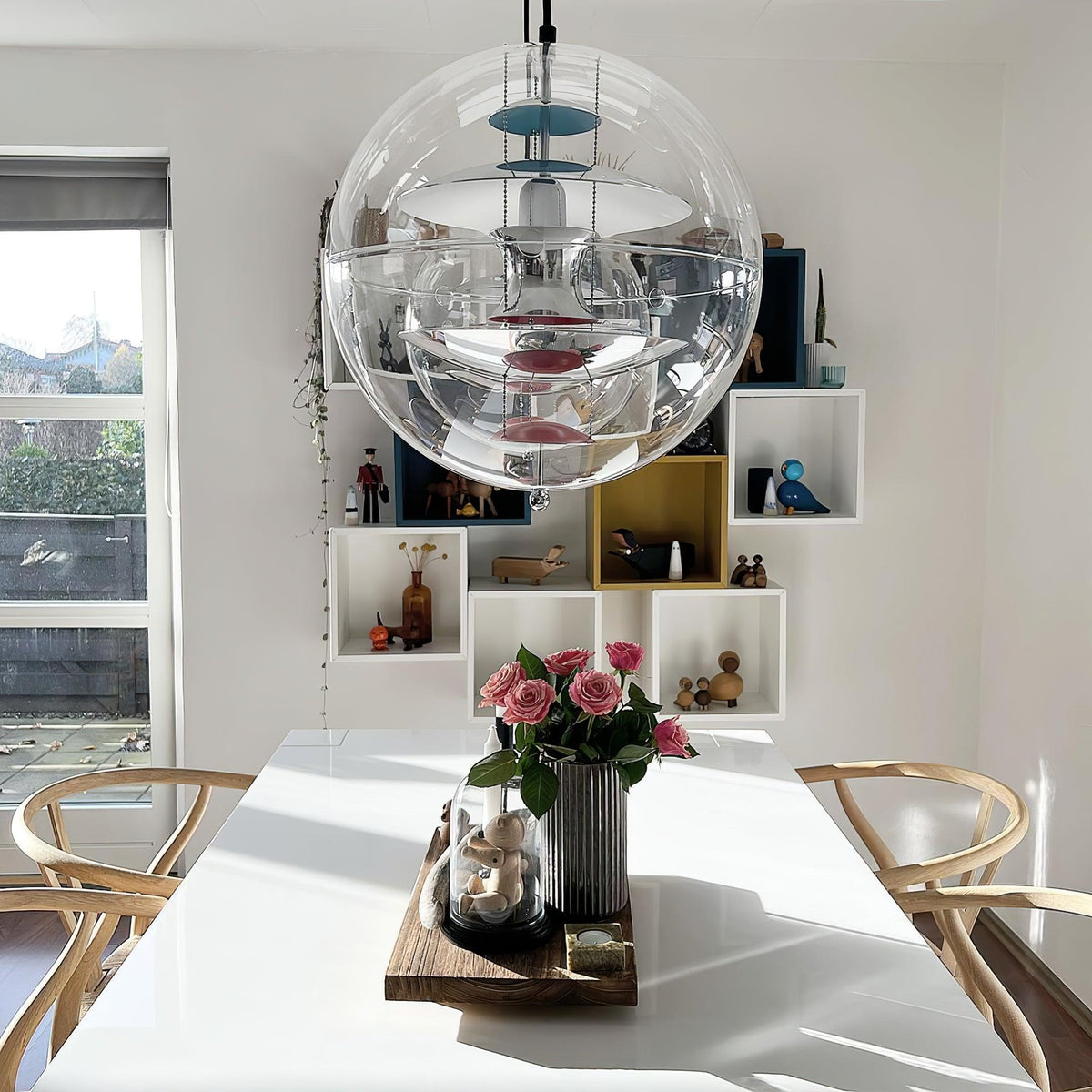 From a low angle, this Clérachic sphere pendant hangs above a white table with pink roses, in front of a window and cube shelves.