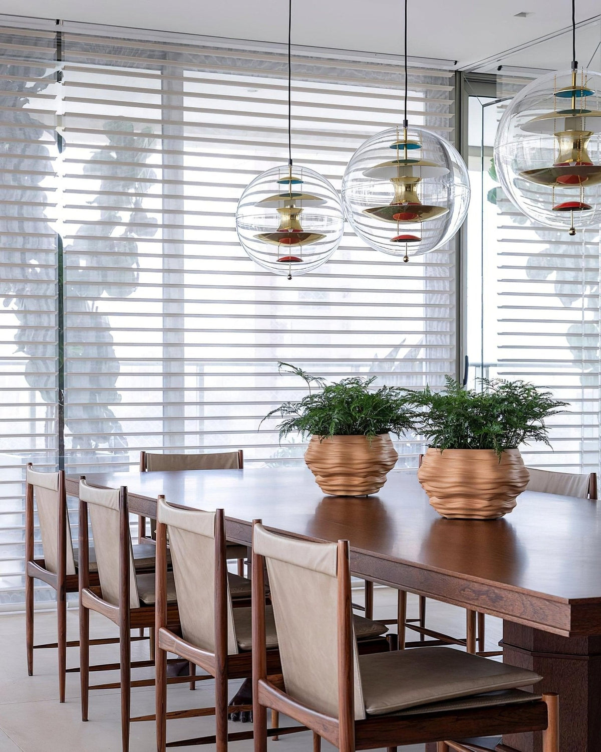 Gold-toned Clérachic sphere pendants in a row over a long wood dining table, with leather-backed chairs and copper planters beneath.