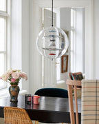 A Clérachic sphere pendant hangs by tall windows over a dark table with a patterned vase of pale flowers and a rattan chair.